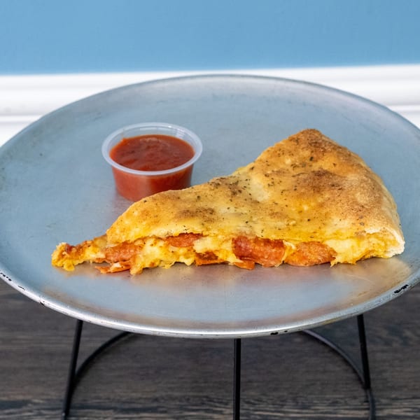 A slice of pepperoni pizza calzone with a side of marinara sauce on a metal tray.